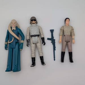 May include: Three plastic action figures and one blue rifle accessory. The first figure wears a long teal robe with a tan headpiece. The second figure wears a light gray uniform with a helmet and black boots. The third figure wears a tan top and pants.
