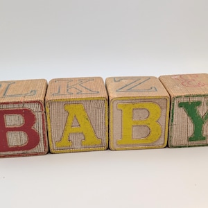 May include: Four vintage wooden blocks spelling "BABY". The blocks are light brown with red, yellow, and green letters. The blocks have a worn, aged appearance, suggesting they are antique toys.