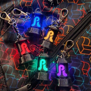 May include: A collection of black lantern-shaped keychains with a glowing "R" design in various colors, including blue, red, green, yellow, and purple. Each keychain has a silver clasp. The keychains are on a patterned black surface.