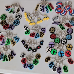 May include: A collection of colorful keychains with various designs, including geometric shapes, symbols, and text. The keychains feature a mix of colors like red, blue, green, and yellow, with silver key rings.