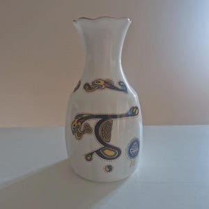 Könnte beinhalten: Eine weiße Keramikvase mit gewelltem Rand, die auf dem Körper ein dekoratives, keltisch inspiriertes Knotenmuster in Schwarz, Gelb und Blau aufweist. Ein kleiner, runder blau-weißer Aufkleber ist an der unteren rechten Seite der Vase angebracht.