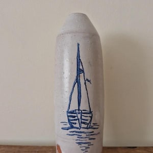 Peut inclure: Un vase cylindrique en céramique avec un émail blanc cassé moucheté, orné d'une illustration de voilier bleu dessinée à la main sur le devant. La base du vase est en terre cuite non émaillée avec une petite marque circulaire visible.