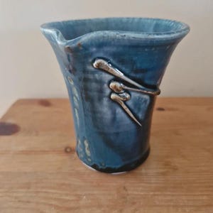 Può includere: Una brocca in ceramica blu con finitura lucida e bordo svasato dotato di un piccolo beccuccio. La superficie esterna presenta uno smalto blu intenso con sottili variazioni di tono e un motivo lineare bianco in rilievo sul lato.