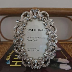 Può includere: Una cornice portafoto ovale in metallo color argento con un intricato design paisley traforato impreziosito da strass trasparenti. L'inserto della cornice riporta il testo: PAST TIMES, Set of Three Diamante Paisley Frames, 7.7cm x 5.1cm.