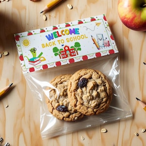 Puede incluir: Dos galletas de avena y pasas en una bolsa de plástico transparente con un encabezado "WELCOME BACK TO SCHOOL". El encabezado tiene ilustraciones con temas escolares. Lápices y manzanas están esparcidos alrededor de las galletas.