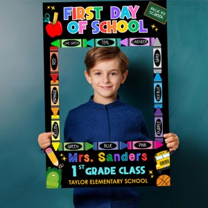 Pode incluir: Uma moldura preta com o texto "FIRST DAY OF SCHOOL" em letras coloridas. A moldura é decorada com lápis de cor, uma maçã, um autocarro escolar e uma bola de basquete. A moldura diz "Mrs. Sanders 1st Grade Class". Um menino segura a moldura.