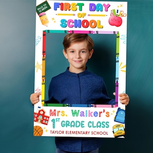May include: A white photo booth prop with the text "FIRST DAY OF SCHOOL" in colorful letters. The prop features school-themed illustrations like a pencil, apple, and school bus. A young boy is holding the prop, smiling at the camera.