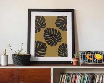 May include: A framed wall art print featuring a repeating pattern of dark charcoal monstera leaves set against a solid mustard gold background. The artwork is enclosed in a simple black frame with a wide white mat border, creating a clean contrast.
