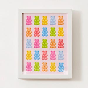 May include: A framed print with a pink background featuring a grid of 25 gummy bear shaped like teddy bears in various colors, including pink, blue, green, yellow, and orange.