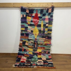 May include: A rectangular, handmade rug with a vibrant, geometric design. The rug features a patchwork of colours including red, yellow, blue, green, and purple. The rug is hanging against a white wall and is approximately 152 cm long.