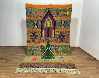 May include: A colorful, hand-woven rug with a geometric design. The rug features a central archway motif in shades of red, yellow, and purple, with a green tree-like shape below. The background includes orange, brown, and green sections with decorative patterns.