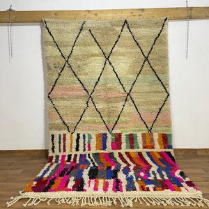 Pode incluir: Um tapete marroquino bege e multicolorido com um padrão de losangos e uma parte inferior vibrante com franjas. O tapete apresenta formas de losangos pretos, listras rosa e verde e um design geométrico colorido em tons de rosa, azul e laranja.