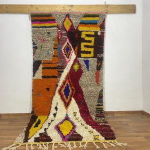 Puede incluir: Una colorida alfombra geométrica con una mezcla de texturas y patrones. La alfombra presenta un diseño central crema con formas de diamante en azul, amarillo y rosa. Tiene secciones naranjas, marrones y amarillas, con flecos en la parte inferior.