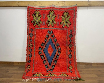Puede incluir: Una vibrante alfombra tejida a mano con un fondo rojo intenso. La alfombra presenta un diseño central de diamante azul, enmarcado por un borde de formas geométricas más pequeñas en varios colores. La parte superior tiene tres patrones florales.