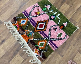 May include: A small, colorful, hand-woven rug with a geometric pattern. The rug features a pink central field with green and purple accents. The design includes diamond and square shapes in orange, blue, and red. The rug has a fringed edge.