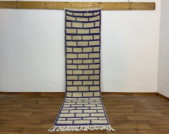 May include: A rectangular rug with a brick-like pattern. The rug features a beige background with dark blue rectangular shapes, resembling bricks. The rug has a fringe at the bottom and is displayed on a wooden floor.