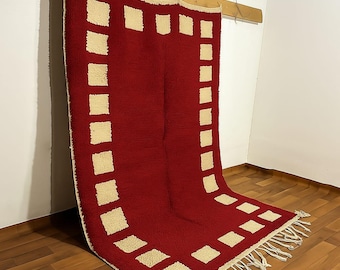 Könnte beinhalten: Ein roter Teppich mit einem cremefarbenen quadratischen Muster. Der Teppich hängt an einem Holzbalken. Der Teppich hat einen Fransen am unteren Rand. Der Teppich besteht aus einem dicken, strukturierten Material.