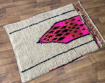 May include: Rectangular rug with a cream-colored background and a vibrant pink geometric design. The rug features black stripes and a colorful fringe. A decorative piece for home decor.