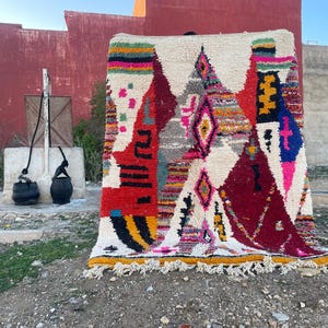 May include: A colorful Moroccan rug with geometric patterns in shades of red, pink, yellow, blue, and white. The rug is made of wool and has a fringe border.