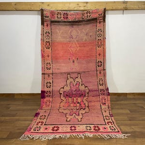 May include: A rectangular, hand-woven rug with a pink and peach color scheme. The rug features geometric patterns and a central floral design. The edges are bordered with a dark brown trim and a fringe.
