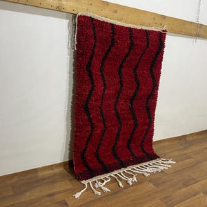 Könnte beinhalten: Ein roter und schwarzer Teppich mit Zickzackmuster und zotteliger Textur. Der Teppich hat einen weißen Fransen am unteren Rand und hängt an einem Holzbalken. Der Teppich ist ein dekorativer Gegenstand.