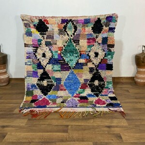 May include: A colorful, handmade rug with a geometric diamond pattern. The rug features a variety of colors including blue, green, pink, and black. The rug is made of a soft, textured material and has a fringed edge. This rug is perfect for adding a pop of color to any room.