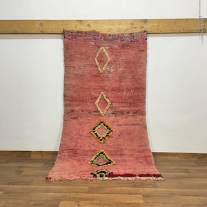 May include: A rectangular, pink Moroccan rug with diamond patterns in yellow, black, and green. The rug is hanging on a wooden beam against a white wall and a wooden floor.