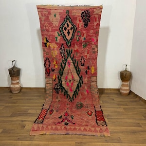 Puede incluir: Una alfombra larga y rectangular de color rojo con patrones geométricos en verde, negro, amarillo y rosa. La alfombra tiene un diseño central en forma de diamante y un borde con flecos. La alfombra se exhibe sobre un suelo de madera.