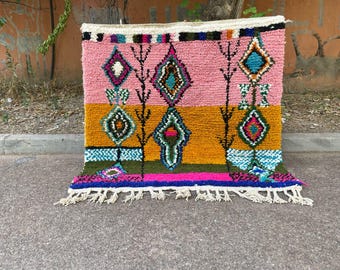 May include: A colorful, rectangular rug with a geometric design. The rug features a pink top section, a yellow-orange middle section, and a green and blue bottom section. The design includes diamond and tree-like shapes in various colors. White fringe lines the bottom edge.