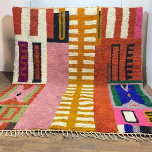 May include: A rectangular area rug featuring a high-pile texture and a vibrant geometric pattern. The design includes blocks of orange, pink, cream, yellow, blue, and green, accented with black linear motifs and a fringed edge at the bottom.