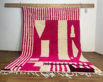 May include: A vibrant, hand-woven rug in shades of pink and cream. The rug features a bold design with large, stylized letters and a striped border. The rug has a fringed edge and is displayed against a wooden background.