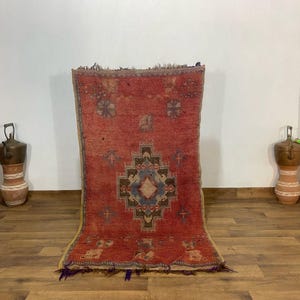 May include: A rectangular, red rug with a central geometric design in brown, blue, and cream. The rug has a distressed appearance with visible wear and tear, and a fringe along the bottom edge. Two decorative vases are on either side.
