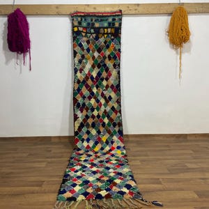 May include: A long, colorful rug with a diamond pattern in various colors, including red, blue, green, and yellow. The rug has a fringed edge and is displayed against a white wall. Yarn bundles hang above.