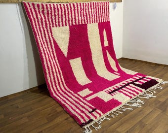 May include: A vibrant pink and cream-colored rug with a bold geometric design. The rug features large cream-colored letter shapes against a pink background, with vertical striped borders and fringe detailing. The rug is made of a thick, textured material.