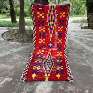 Pode incluir: Um tapete retangular vibrante com fundo vermelho e padrões geométricos em amarelo, azul e rosa. O tapete é exibido ao ar livre, mostrando seu design intrincado e paleta de cores. As dimensões do tapete não são visíveis.