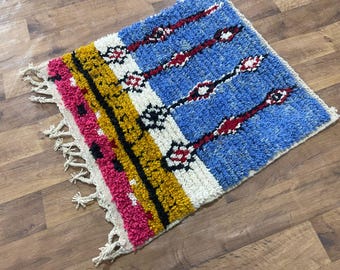 May include: A small, rectangular rug with a vibrant design. The rug features a blue field with red and white geometric patterns, and stripes of pink, yellow, black, and white. It has a fringed edge.
