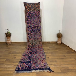 May include: A long, rectangular rug with a diamond pattern in shades of blue, pink, and orange. The rug has a brown background and a fringed edge. The rug is placed on a wooden floor.