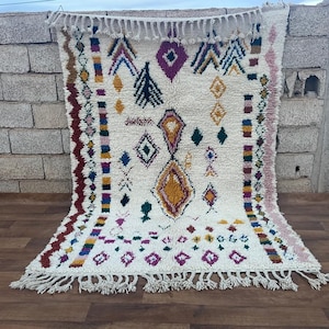 May include: A white Moroccan rug with a geometric pattern in shades of red, pink, yellow, blue, and green. The rug has a fringe border.