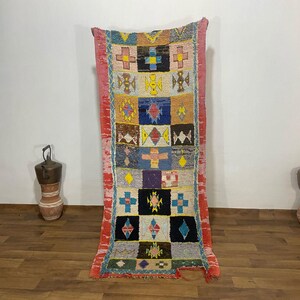 Op de afbeelding: Een rechthoekig, kleurrijk tapijt met een geometrisch patroon. Het tapijt heeft een centraal ontwerp van vierkanten in verschillende kleuren, waaronder blauw, geel en rood. Het tapijt is ongeveer 183 cm lang.