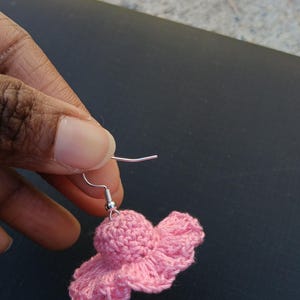 May include: A single pink crochet earring shaped like a small hat. The earring is held up by a silver-coloured hook. The crochet work has a textured appearance, with a ruffled brim. The earring is set against a dark background.