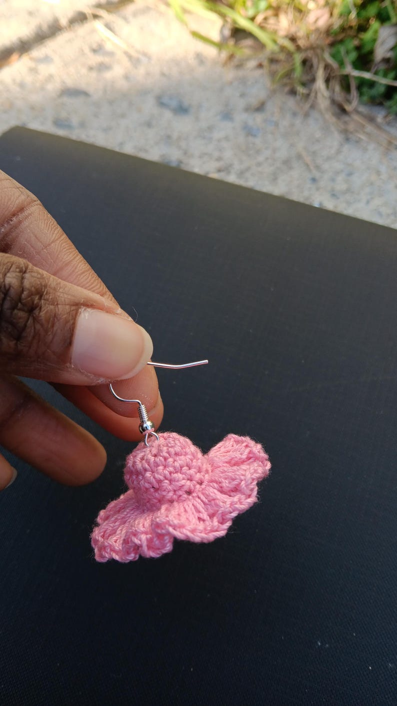 May include: A single pink crochet earring shaped like a small hat. The earring is held up by a silver-coloured hook. The crochet work has a textured appearance, with a ruffled brim. The earring is set against a dark background.