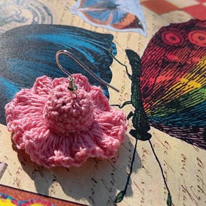 May include: A pink crocheted flower earring with a silver hook. The earring is set against a backdrop featuring butterfly illustrations with vibrant rainbow and blue wings. The background has a vintage aesthetic.