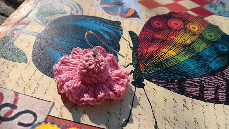 May include: A single pink crocheted flower earring with a silver hook. The earring is set against a backdrop featuring butterfly illustrations with rainbow and blue wings. The background has a vintage aesthetic.