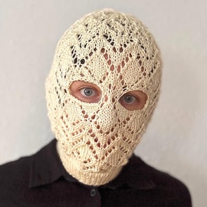 Könnte beinhalten: Cremefarbene Strick-Balaclava mit Lochmuster. Die Sturmhaube bedeckt den gesamten Kopf und Hals, mit Öffnungen für die Augen. Die Person trägt ein schwarzes Hemd mit Knöpfen.