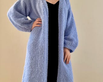 Könnte beinhalten: Langer, hellblauer Cardigan mit weicher, strukturierter Optik. Der Cardigan hat lange Ärmel und eine knielange Form. Die offene Vorderseite zeigt ein schwarzes Kleid darunter. Ein gemütlicher und stilvoller Oberbekleidungsartikel.