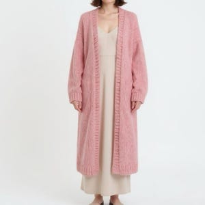 A woman poses in a fluffy, ankle-length pink cardigan and a beige dress.