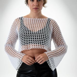 May include: A white, open-weave, long-sleeved top with a boat neck. The top is cropped, revealing the midriff. The model is wearing a black top underneath and black leather pants. The top has a fishnet-like appearance.