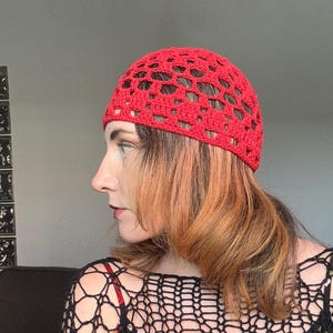 May include: A bright red, open-weave crochet beanie. The hat is made of a thick yarn and has a textured appearance. The beanie is a solid color and is worn on the head.