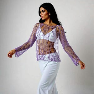 May include: A sheer, lavender crochet top with long sleeves and a round neckline. The open weave design reveals a white undergarment. The top has a scalloped hemline and is paired with light-colored pants.