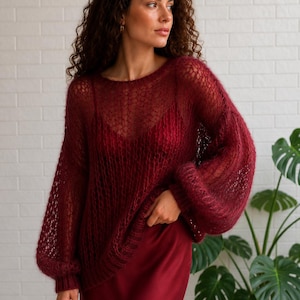 May include: A deep burgundy open-knit jumper with long, voluminous sleeves. The jumper is worn with a matching burgundy satin skirt. The model is standing in front of a white brick wall with a large green plant in the background.